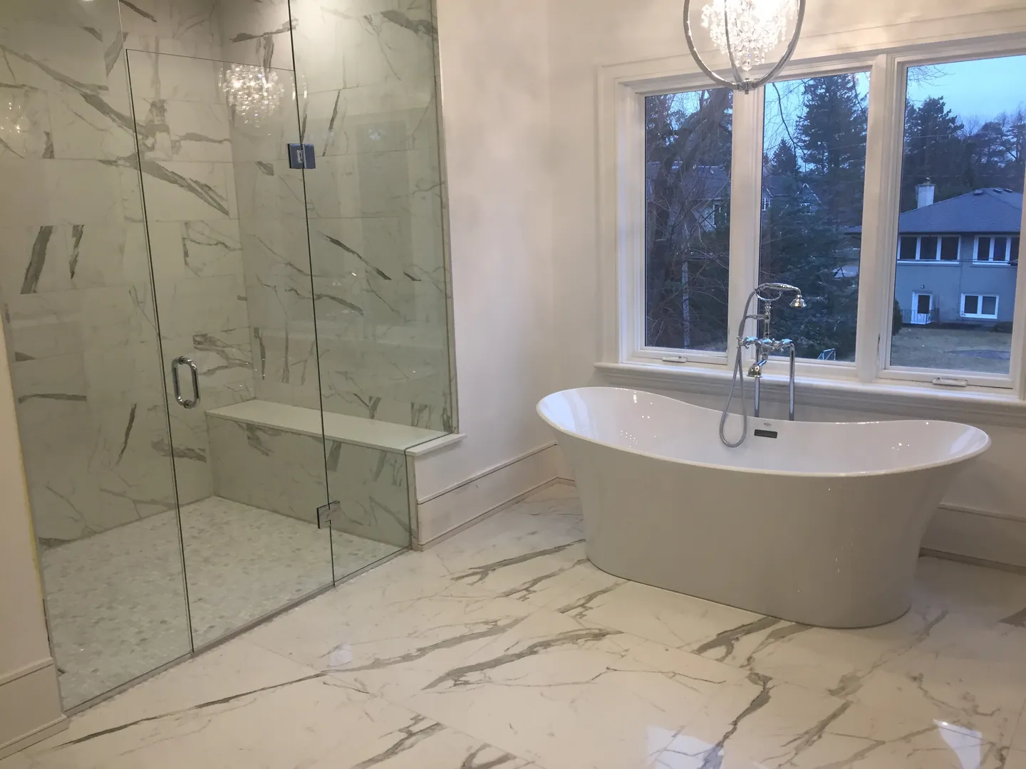 Glass shower and freestanding bath surrounded by pale, veined wall and floor tile
